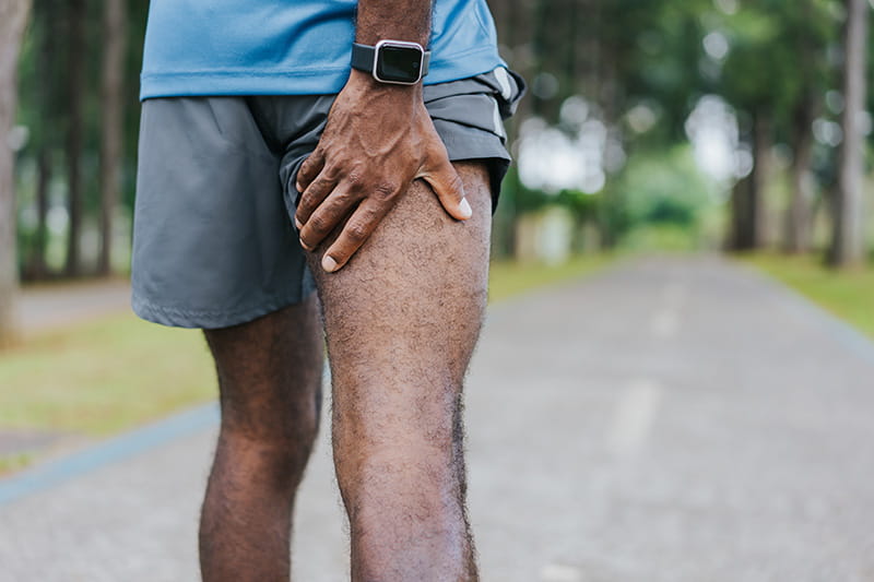 View of a man from the waist down, walking down the road, stopping to grab the back of his leg