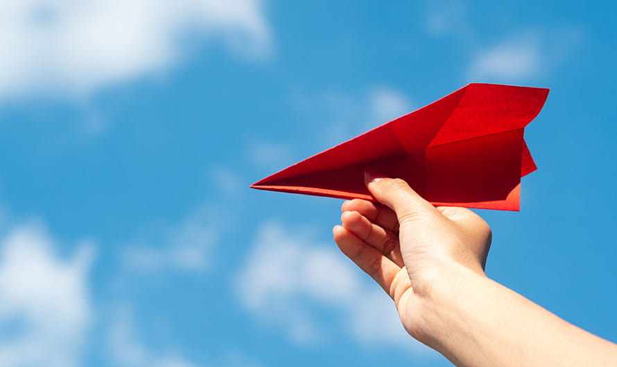A hand holding a red paper airplane against a cloudy blue sky