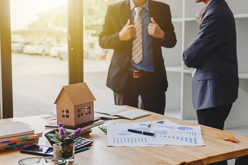 focus on a desk strewn with papers and a small decorative model house while two people stand in the background discussing real estate