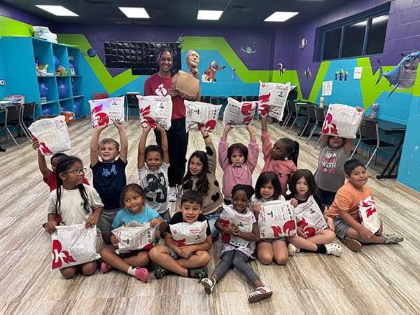an American Heart Association volunteer holding up a CPR manikin, surrounded by a group of children in a classroom